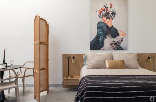 Tomorrow Building, Fully Furnished Apartments, Come With Mid-century Modern Furniture. Boho Room Divider Separates The Dining And Living Area. A Large Painting Of A Person With A Hat Made Of Flowers Hangs Over The Bed Covered In Neutral Prints.