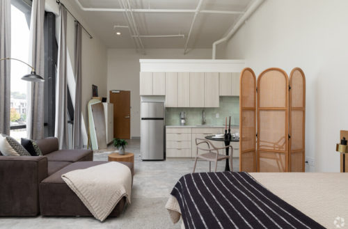 Fully-Furnished Apartments In Downtown Chattanooga. This Tomorrow Building Apartment Showcases Mid Century Modern Furniture, A Medium Sized Fridge, Cozy Couch, And Two Person Dining Table.
